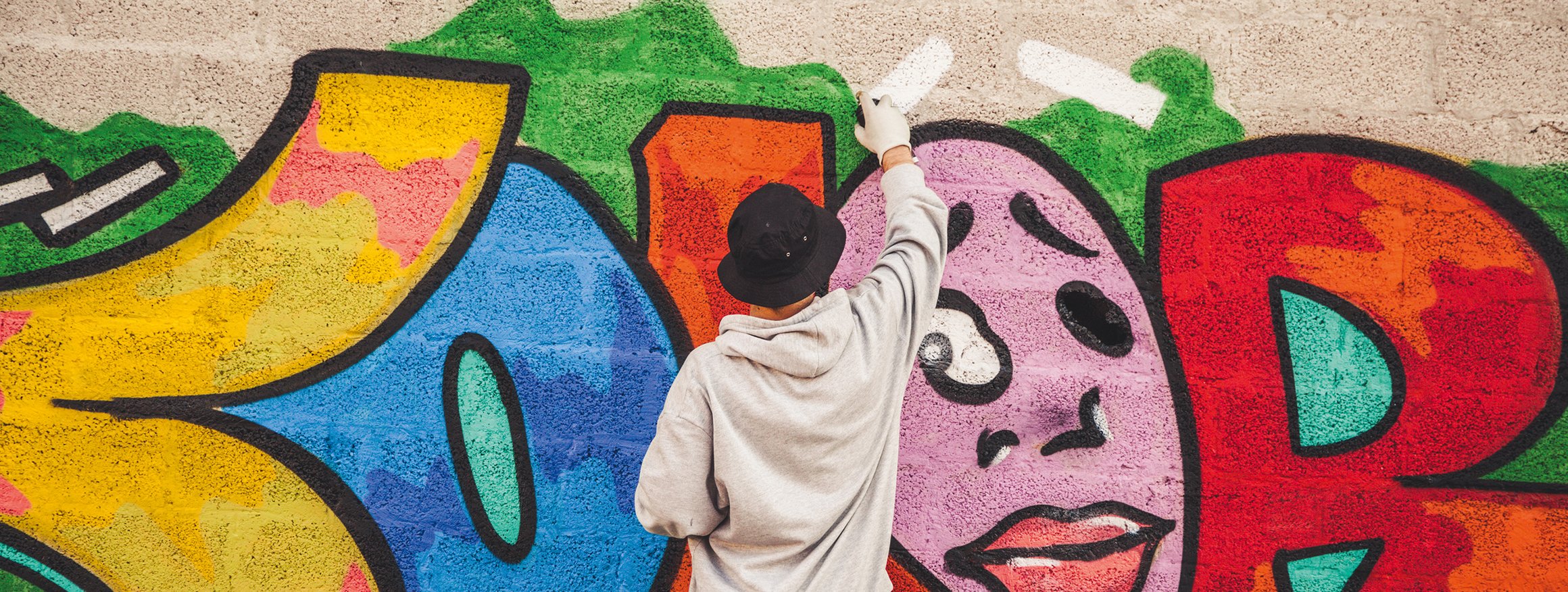 Ein Graffiti-Sprayer mit Mütze und Handschuhen steht an einer Wand und malt mit der Sprühdose ein Bild
