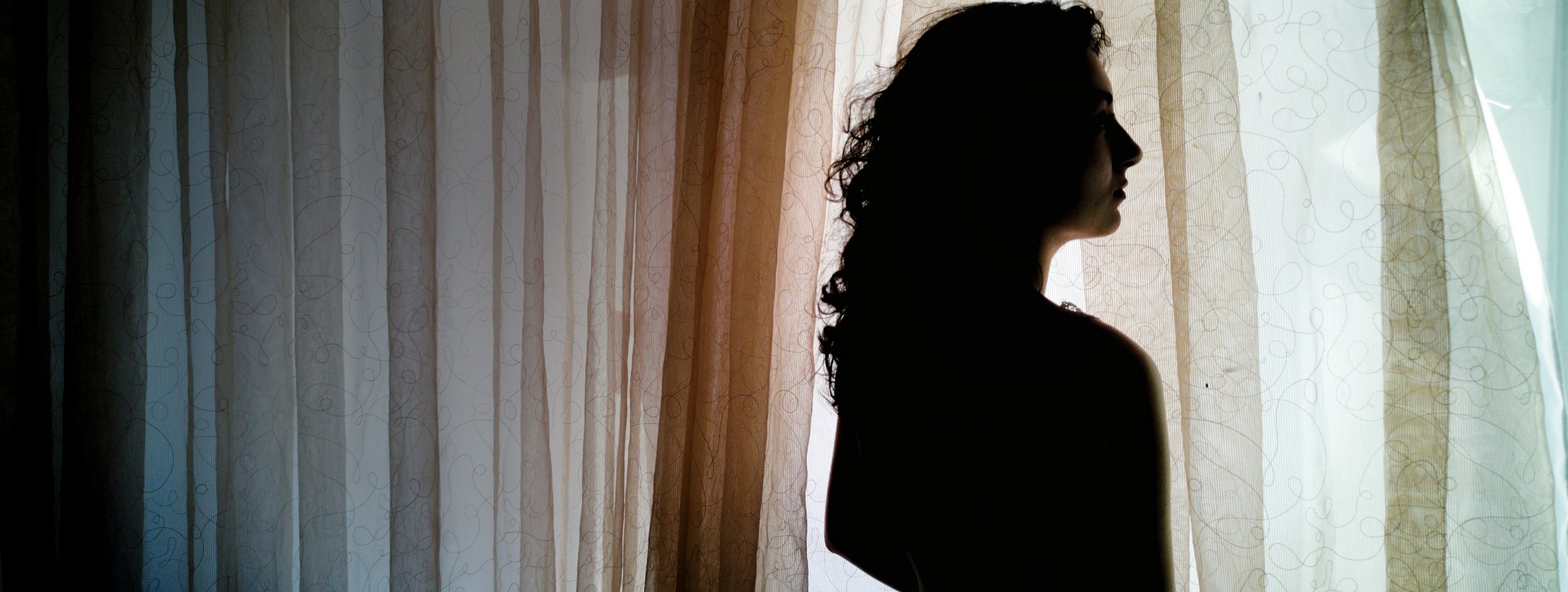 Eine junge Frau steht als dunkle Silhouette am Fenster und schaut hinaus, vor ihr ein weißer Vorhangstoff.