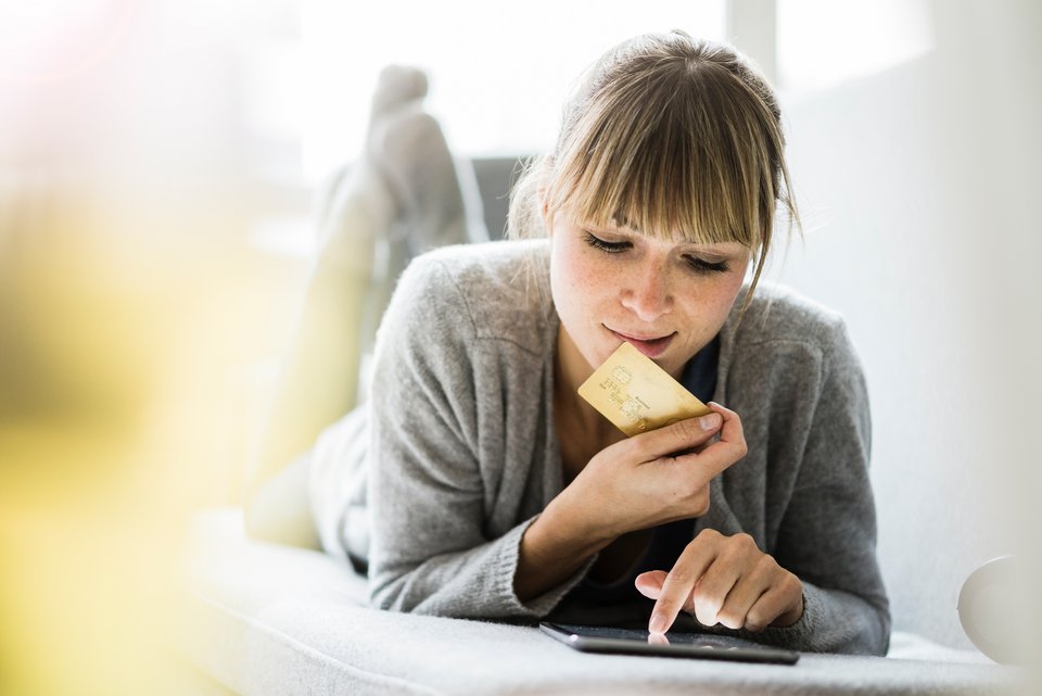 Eine junge Frau liegt auf dem Bauch auf einem Sofa, das Smartphone vor ihr und hält dabei ihre Kreditkarte in der Hand, da sie dem Kaufangebot nicht widerstehen kann