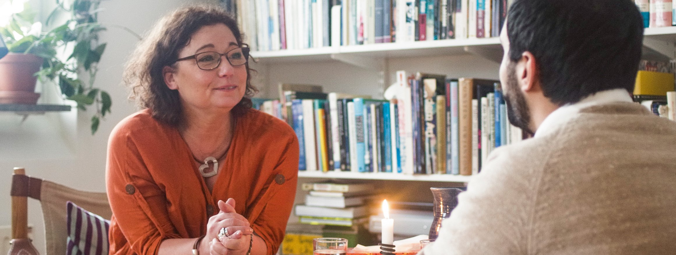 Eine Theapeutin sitzt mit ihrem Patient vor einer Bücherwand, neben ihr ein Beistelltisch mit einer brennenden Kerze