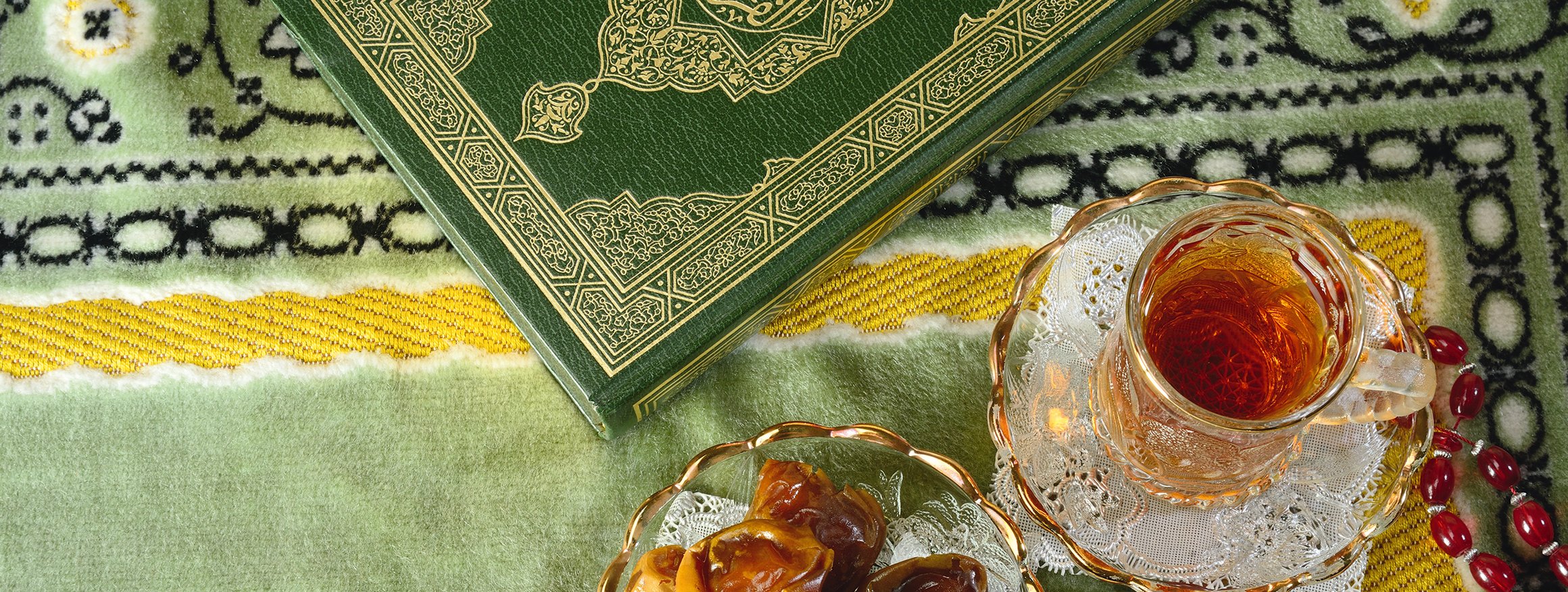 Auf einem grünen orientalischen Teppich liegen der Koran, ein Glas Tee und eine Glasschale mit Datteln