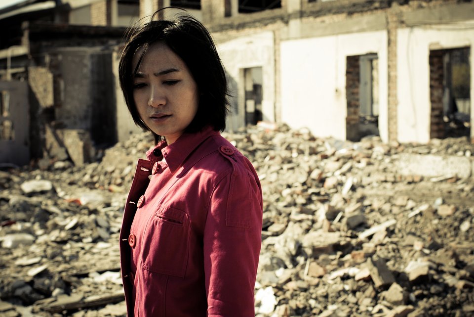 Eine junge Frau mit roter Jacke steht traurig vor einem zertrümmerten Haus