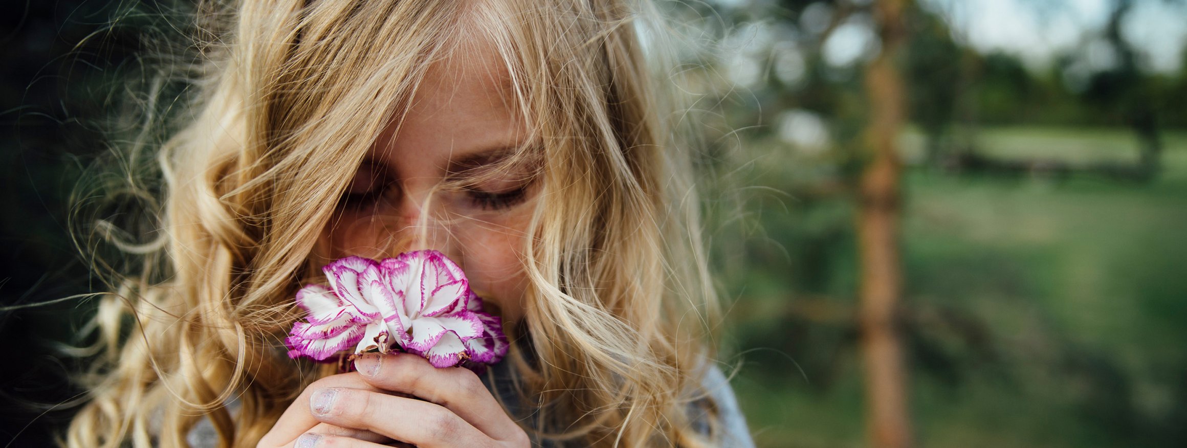 Ein blondes Mädchen mit langen Locken riecht an einer Blume und hat dabei die Augen geschlossen