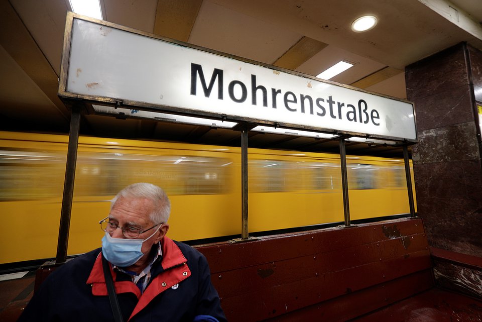 Ein älterer Mann mit Coronamaske sitzt an einer Bahnstation, die Mohrenstraße heißt, während hinter ihm eine Bahn vorbeifährt
