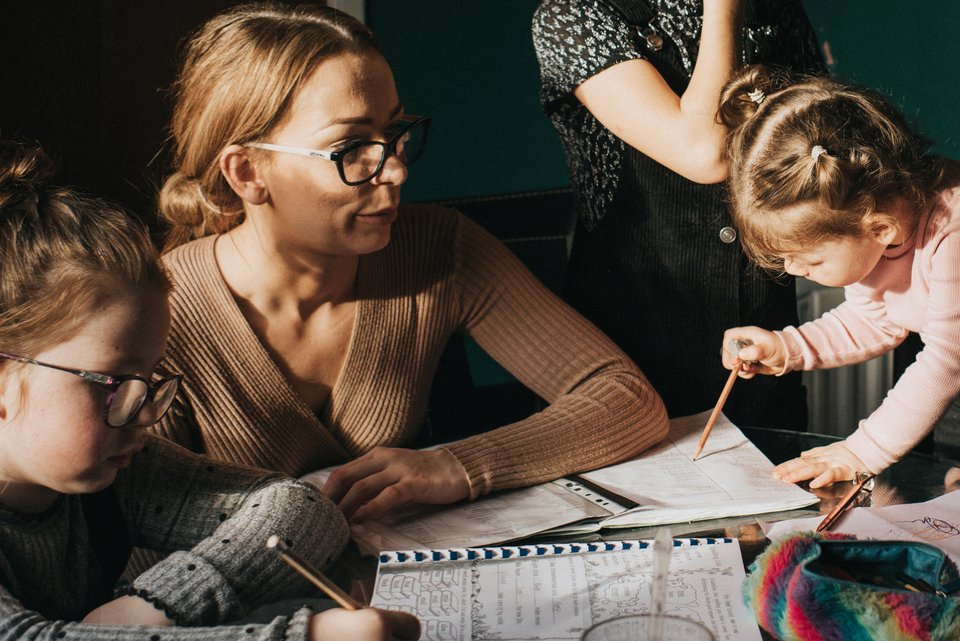 Eine blonde Frau mit Brille sitzt mit ihren Stiefkindern am Tisch und hilft bei den Hausaufgaben