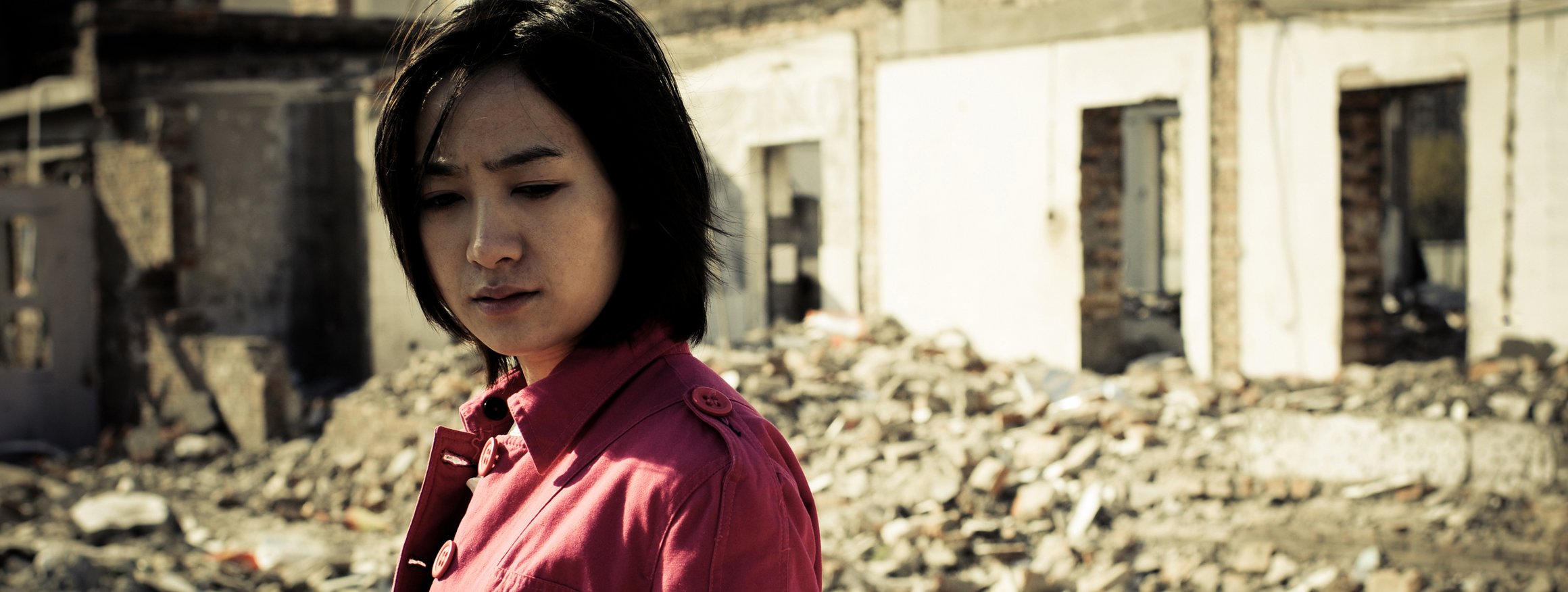 Eine junge Frau mit roter Jacke steht traurig vor einem zertrümmerten Haus