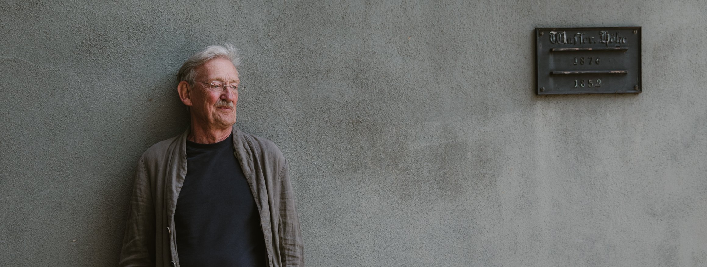 Der Schriftsteller, Christian Haller, lehnt an eine graue Wand mit einer Gedenktafel, die Hände in den Hosentaschen