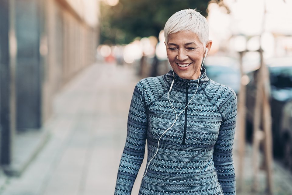 Eine ältere Frau mit grauen kurzen Haaren, läuft in Sportkleidung  und In-Ears lächelnd durch eine Straße