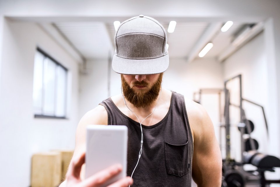 Ein muskulöser Mann im Fitnessstudio mit Baseballkappe und Bart macht mit seinem Smartphone ein Selfie 