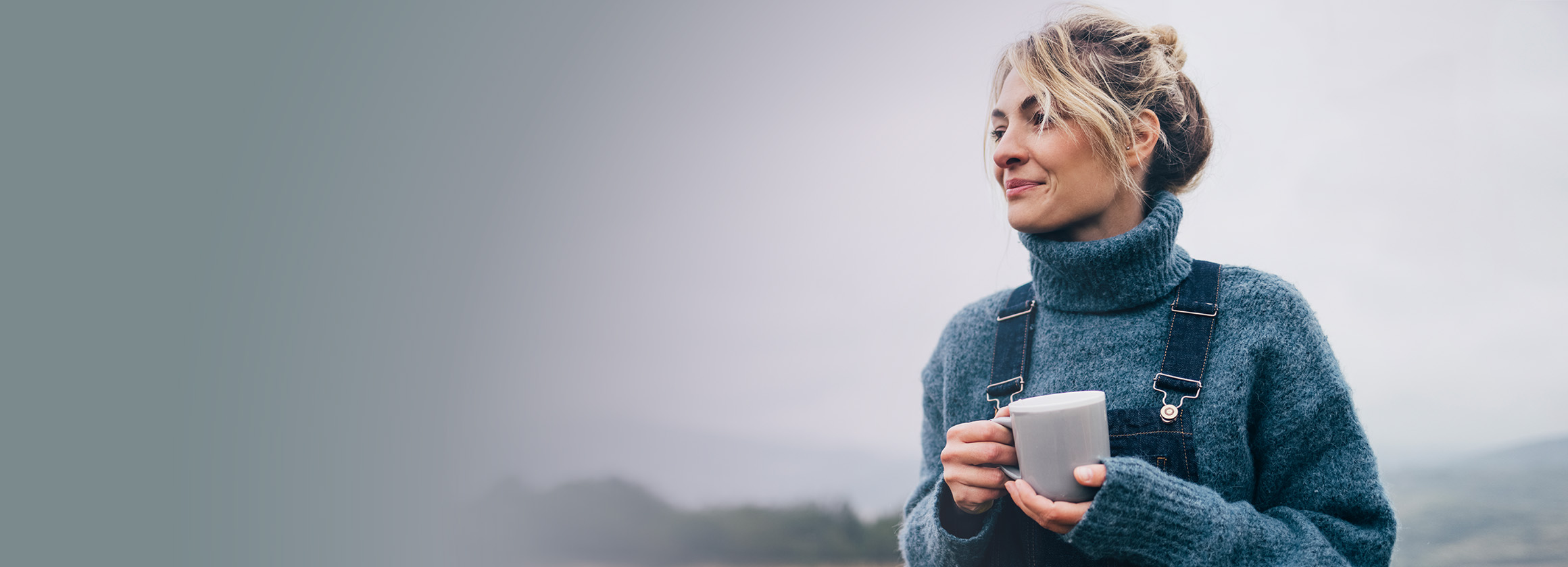 Eine Frau mit blonden Haaren, Latzhose und Pullover, hält einen Tee in der Hand und schaut zufrieden in die Ferne.