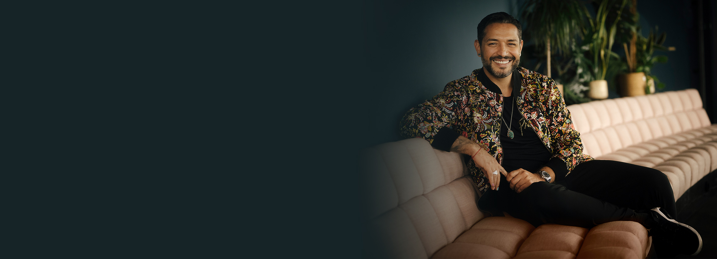 Das Foto zeigt Gianni Jovanovic, wie er auf einem hellen Sofa sitzt. Der Mann lächelt freundlich.